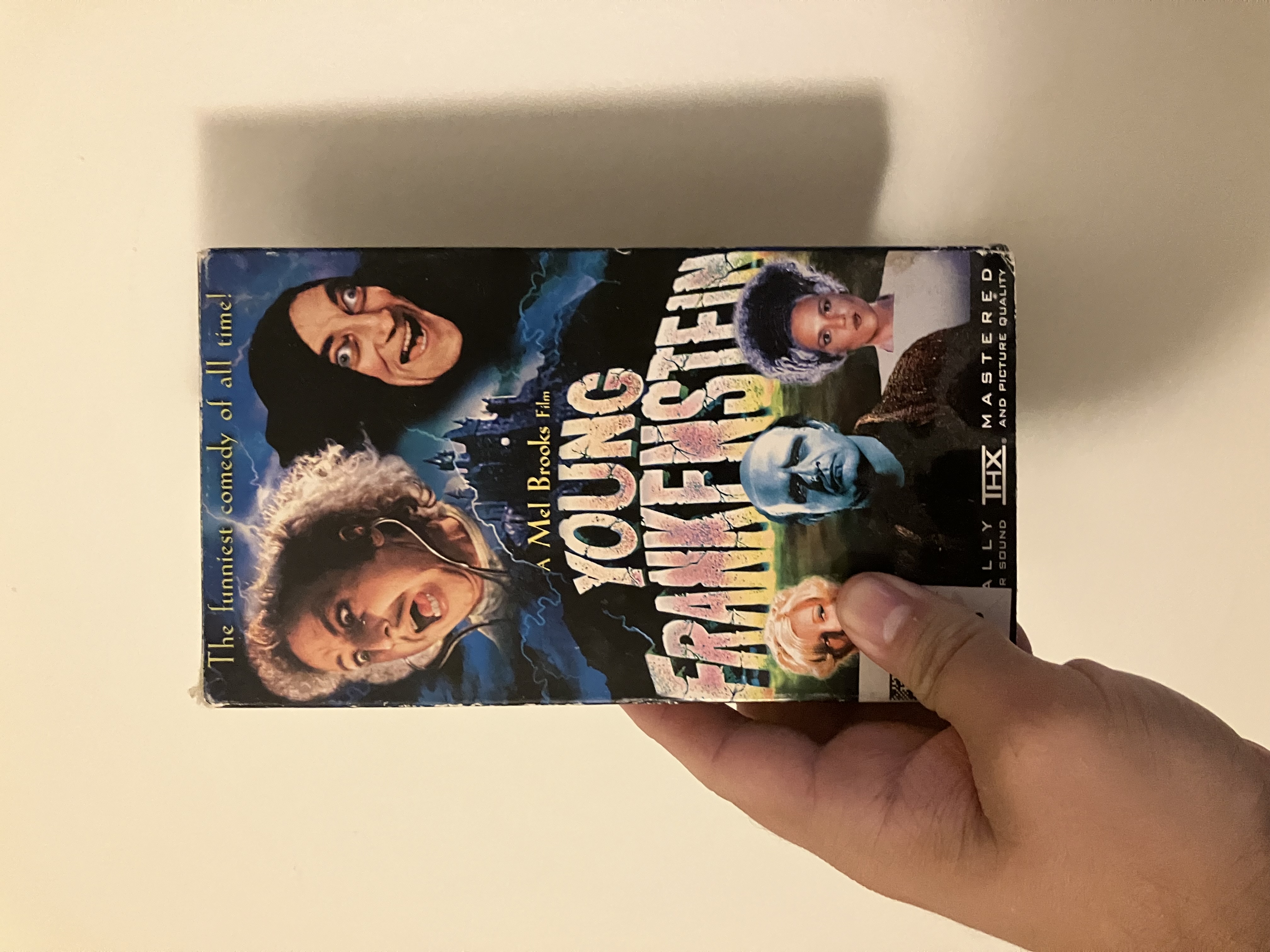 A person holds a VHS tape cover for the film ‘Young Frankenstein.’ The cover features a collage with four characters’ faces: a man with frizzy hair and an expression of surprise, a grinning man in a black hood, a stern-faced greenish-blue Frankenstein-like character in the center, and two women on either side, one with blonde hair and one with an elaborate coiffure. The title ‘Young Frankenstein’ is prominently displayed in stylized letters across the middle of the cover.