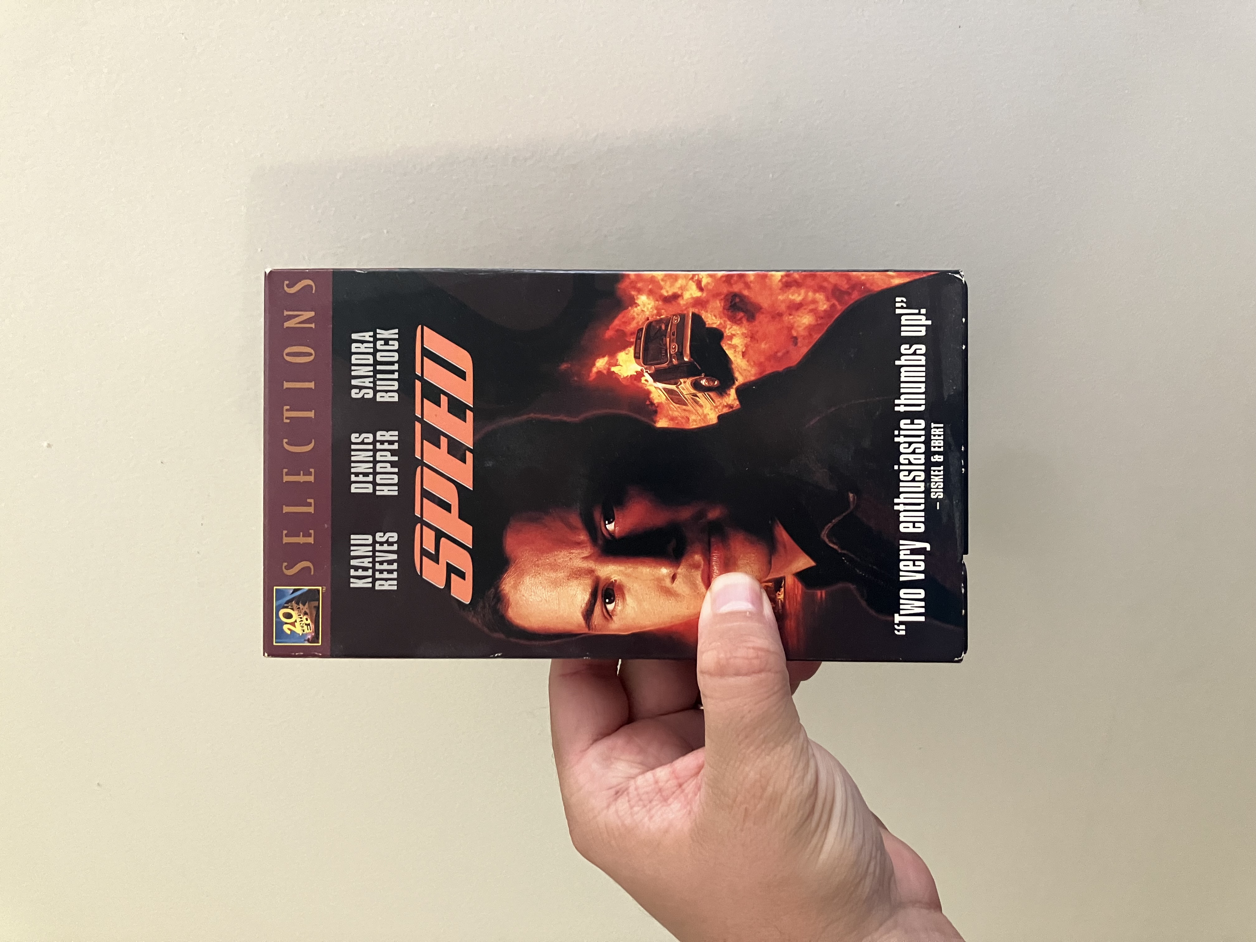 A person holds a VHS tape of the 1994 action film Speed. The cover features a large portrait of Keanu Reeves with a fiery explosion in the background and a bus mid-air. The top lists the stars—Keanu Reeves, Dennis Hopper, and Sandra Bullock—while the movie title Speed appears in bold orange letters. At the bottom is a quote: “Two very enthusiastic thumbs up!” – Siskel & Ebert.